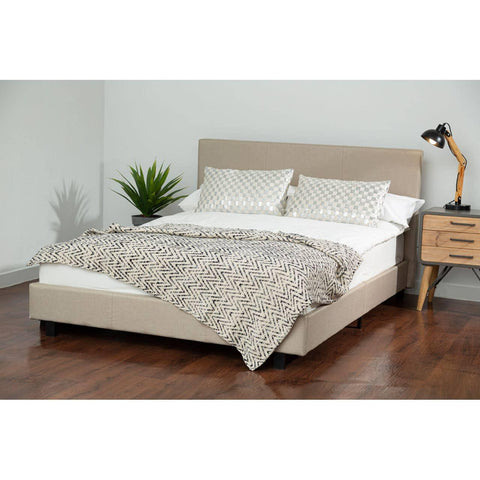 Beige upholstered bed frame with headboard, zigzag throw blanket, pillows, and bedside table