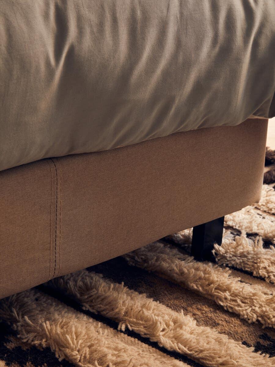 Fabric bed base corner with grey bedding on textured beige and brown rug
