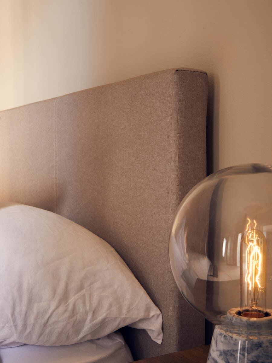 Beige upholstered bed headboard with white pillow and marble table lamp near bedside