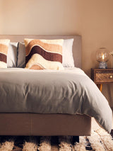 Beige upholstered bed with grey bedding and patterned cushions, bedside table with lamp
