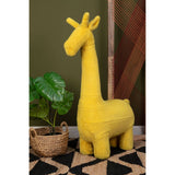 A bright yellow giraffe-shaped stool designed for kids, featuring a tall neck and playful facial features. The stool is positioned next to a green leafy plant and set against a backdrop of earthy tones and geometric patterned flooring