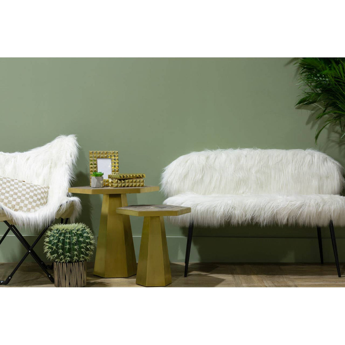 A stylish two-seater sofa with a plush faux fur upholstery in white, featuring sleek black legs. The sofa is positioned next to a modern accent chair and two gold side tables, with a green wall and a potted plant in the background, creating a cozy and contemporary living space