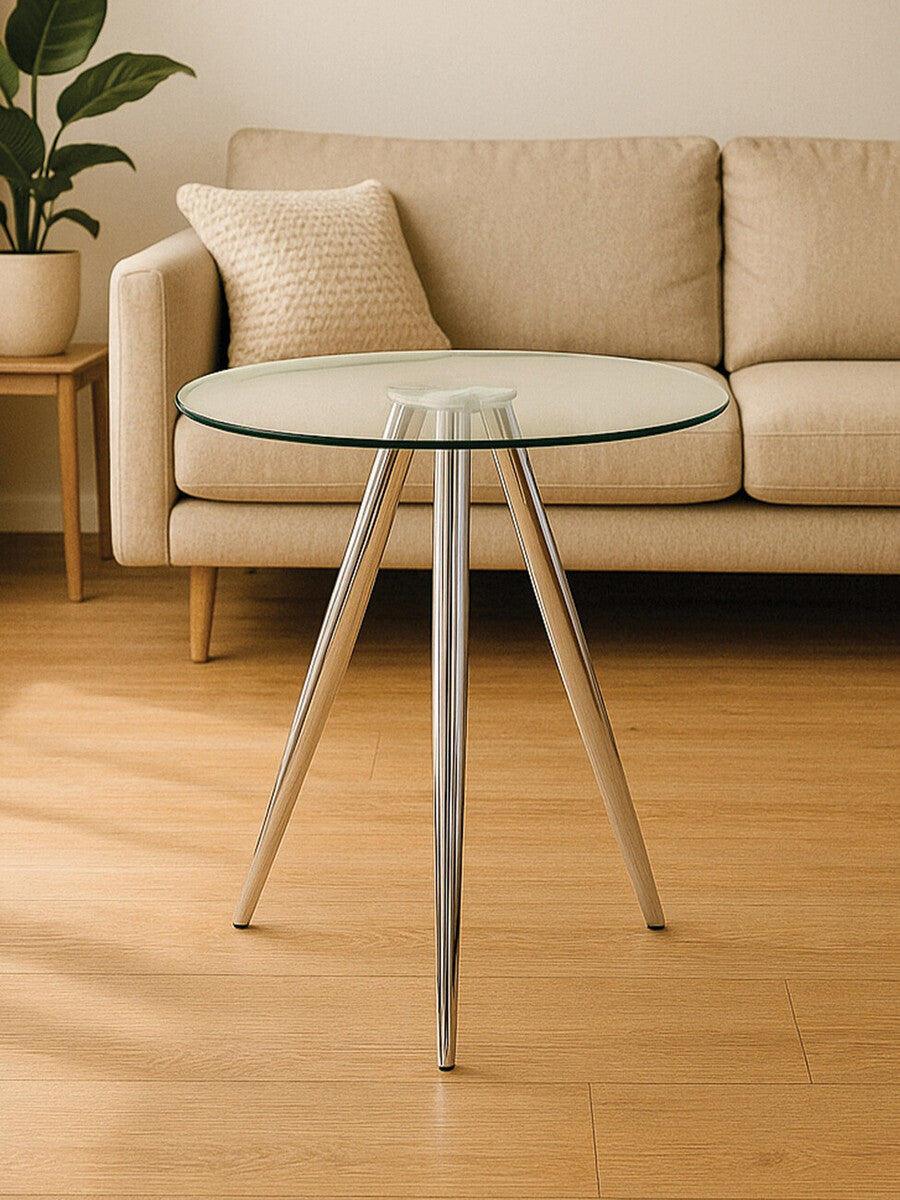 Round glass bar table with chrome legs, featuring a sleek and modern design, positioned on a wooden floor in a well-lit room with a light-colored sofa in the background