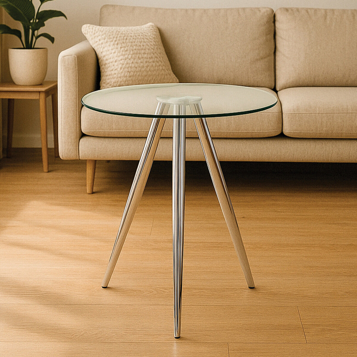 Round glass bar table with sleek chrome legs, positioned in a cozy living space next to a light-colored sofa. The table features a clear glass top and a modern design, complemented by warm wooden flooring and a potted plant in the background
