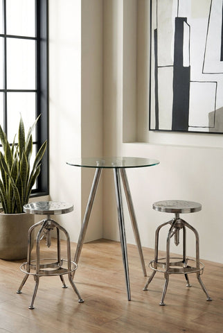 Round glass bar table with chrome legs positioned between two matching bar stools, set against a backdrop of large windows and a mirror, with a potted plant nearby on a wooden floor