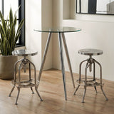 Round glass bar table with sleek chrome legs, accompanied by two matching chrome bar stools, set against a light wooden floor and a bright window with a potted plant in the background
