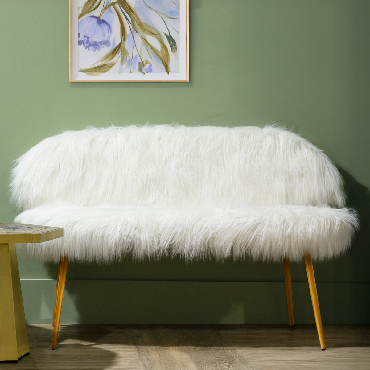 A two-seat sofa covered in plush white faux fur, featuring sleek gold finish legs. The sofa has a rounded backrest and a minimalist design, set against a green wall with a piece of artwork in the background. A small side table is visible next to the sofa