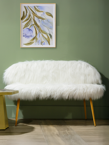 A cozy two-seat sofa upholstered in white faux fur, featuring a rounded back and gold finish legs. The sofa is positioned against a green wall, with a small round side table nearby. A framed artwork with abstract designs hangs above the sofa, adding a touch of color to the scene