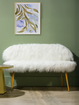 A cozy two-seat sofa upholstered in white faux fur, featuring a rounded back and gold finish legs. The sofa is positioned against a green wall, with a small round side table nearby. A framed artwork with abstract designs hangs above the sofa, adding a touch of color to the scene