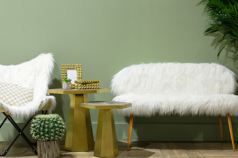Sienna white faux fur two-seat sofa with gold finish legs positioned next to a matching white faux fur chair, accompanied by two gold accent tables. The background features a soft green wall and a potted plant, creating a cozy and modern living space
