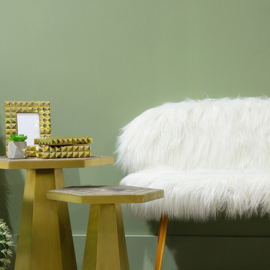 A cozy two-seat sofa upholstered in white faux fur, featuring sleek gold finish legs. The sofa is positioned against a soft green wall, accompanied by two modern gold side tables, one holding a small plant and a decorative frame