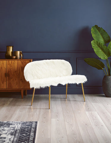 Sienna white faux fur two-seat sofa with a curved backrest and gold finish legs, positioned against a dark blue wall. A wooden sideboard with decorative items is visible in the background, along with a large green plant and a patterned rug on the floor