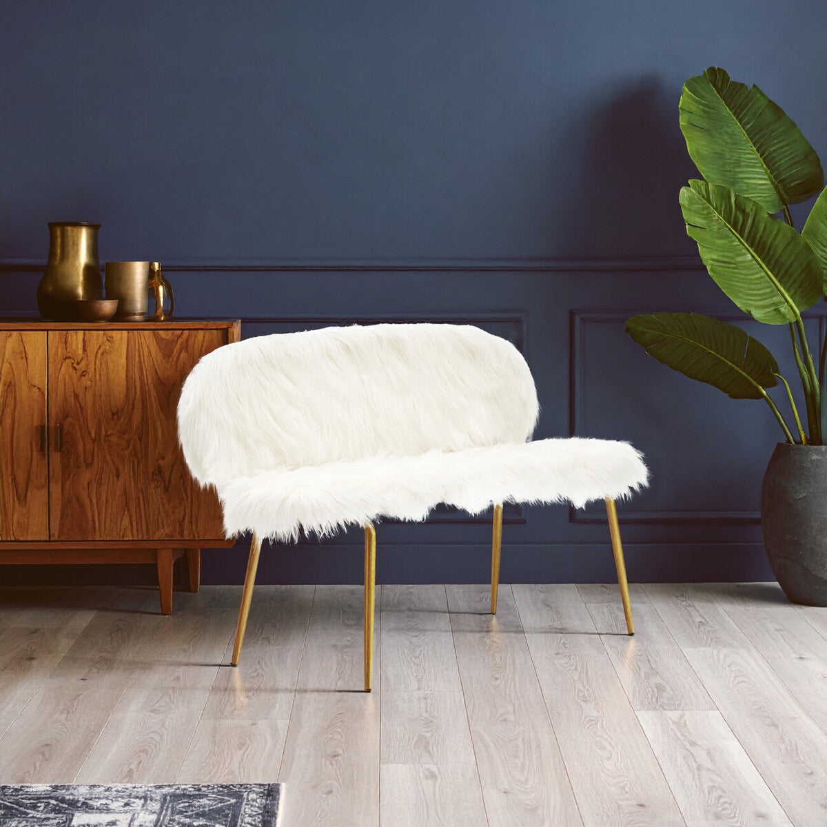 A stylish two-seat sofa featuring a plush white faux fur upholstery and elegant gold finish legs, positioned against a dark blue wall. A wooden side table with decorative items is visible in the background, along with a large green plant in a black pot. The flooring is light-colored wood, enhancing the modern aesthetic of the space
