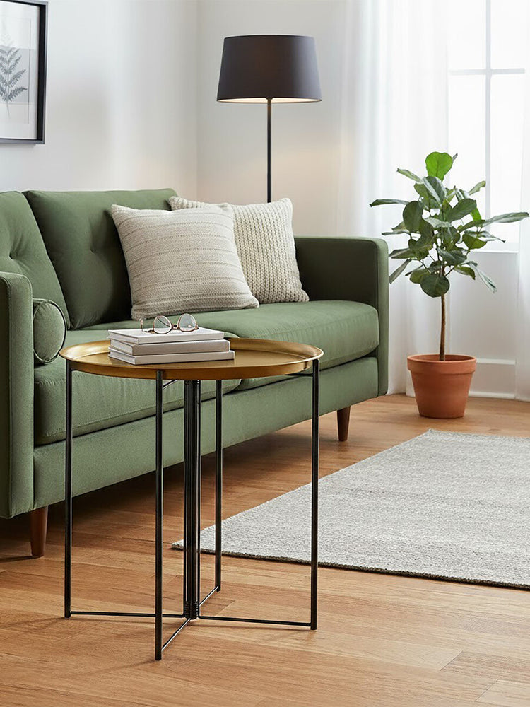 A large side table with a brass top and black metal legs positioned next to a green sofa with two cream-colored pillows. A floor lamp stands nearby, and a potted plant adds greenery to the scene. The flooring is wooden, and a light-colored rug is visible beneath the table. Natural light filters through a window in the background