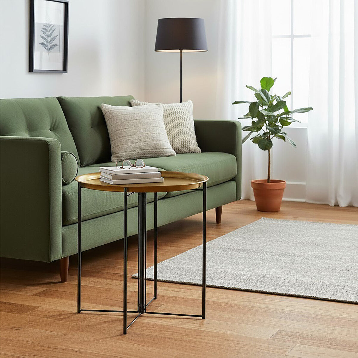 A large side table with a brass top and black metal legs positioned next to a green sofa adorned with decorative pillows. A floor lamp and a potted plant are visible in the background, along with a light-colored area rug on a wooden floor