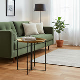 A large side table with a brass top and black metal legs positioned next to a green sofa adorned with decorative pillows. A floor lamp and a potted plant are visible in the background, along with a light-colored area rug on a wooden floor