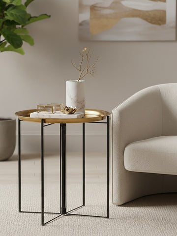 A stylish side table featuring a round wooden top with a brass finish and a black metal frame. The table is positioned next to a light-colored, modern sofa and a decorative plant in the background, creating a cozy living space ambiance. A white candle and a small stack of books are placed on the table's surface