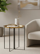 A stylish side table featuring a round wooden top with a brass finish and a black metal frame. The table is positioned next to a light-colored, modern sofa and a decorative plant in the background, creating a cozy living space ambiance. A white candle and a small stack of books are placed on the table's surface