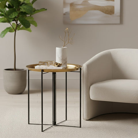 A large side table featuring a brass and black finish, with a wooden top and sleek metal legs, positioned next to a light-colored armchair and a potted plant in a modern living space. A white vase and decorative items are placed on the table's surface