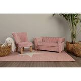 A pink velvet kids chair with a tufted backrest is positioned next to a matching pink sofa on a light-colored rug featuring a star design. A cozy throw blanket is draped over the arm of the sofa, and two woven baskets are placed nearby, adding a warm touch to the room's decor. A green plant is visible in the background against a neutral wall