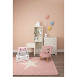 A pink velvet kids chair with wooden legs is positioned in a softly lit room featuring a light pink wall. Nearby, a white dresser and a small white table are visible, along with stacked storage boxes in pastel colors. A star-patterned rug lies on the floor, and colorful balloons are floating in the background