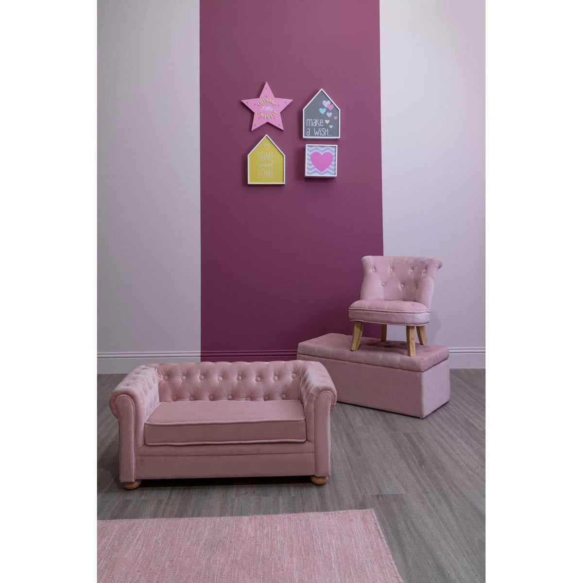 A pink velvet kids chair with a tufted backrest is positioned in a stylish room featuring a purple accent wall. Nearby, a matching pink velvet sofa and a small wooden table are visible, along with colorful wall decorations in the shape of houses and a star. A soft pink rug covers the floor, adding to the cozy atmosphere