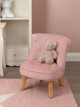 A small pink velvet chair designed for kids, featuring a tufted backrest and wooden legs. A soft teddy bear is placed on the seat, and the background includes a light-colored shelf and a pastel rug. The wall is painted in a soft pink hue