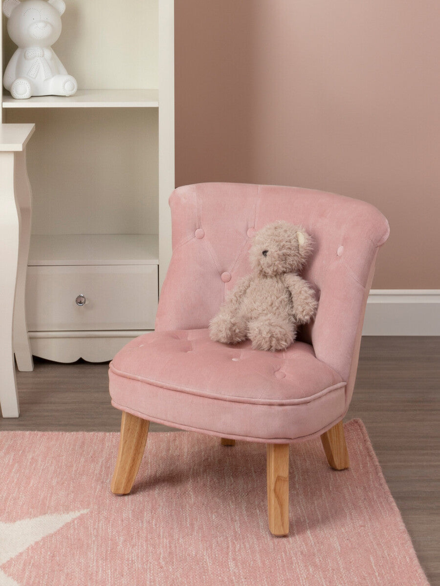 A small pink velvet chair designed for kids, featuring a tufted backrest and wooden legs. A soft teddy bear is placed on the seat, and the background includes a light-colored shelf and a pastel rug. The wall is painted in a soft pink hue