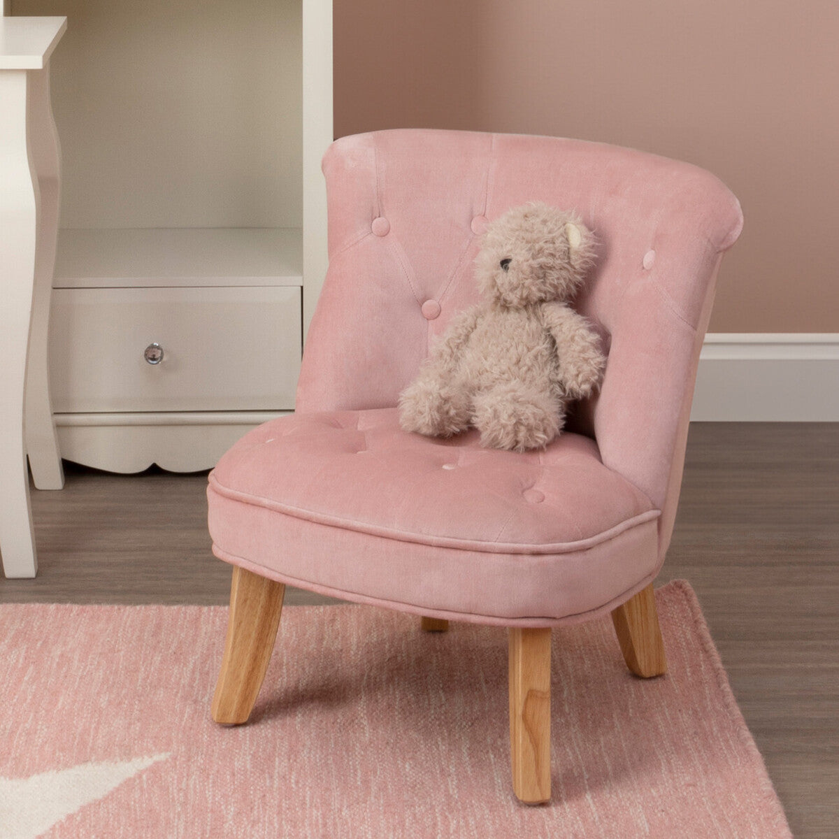 A small pink velvet chair designed for kids, featuring a tufted backrest and wooden legs. A plush teddy bear is seated on the chair, and the background includes a soft rug and a light-colored wall