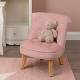 A small pink velvet chair designed for kids, featuring a tufted backrest and wooden legs. A plush teddy bear is seated on the chair, and the background includes a soft rug and a light-colored wall