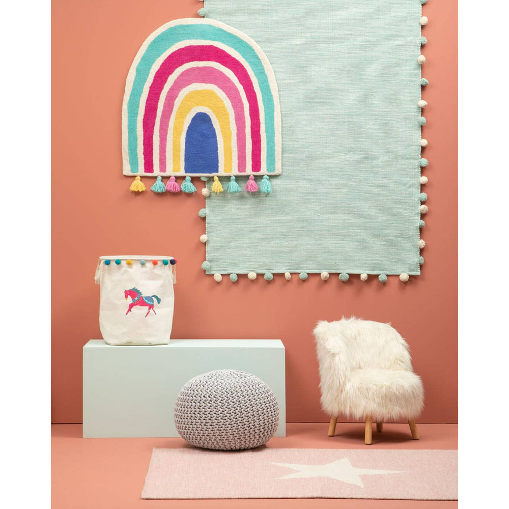A cozy kids' chair covered in white faux fur, featuring wooden legs, positioned in a colorful room with a pink wall and a light blue rug. A playful rainbow wall decoration and a whimsical unicorn storage bin are visible in the background, along with a soft gray pouf