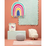 A cozy kids' chair covered in white faux fur, featuring wooden legs, positioned in a colorful room with a pink wall and a light blue rug. A playful rainbow wall decoration and a whimsical unicorn storage bin are visible in the background, along with a soft gray pouf