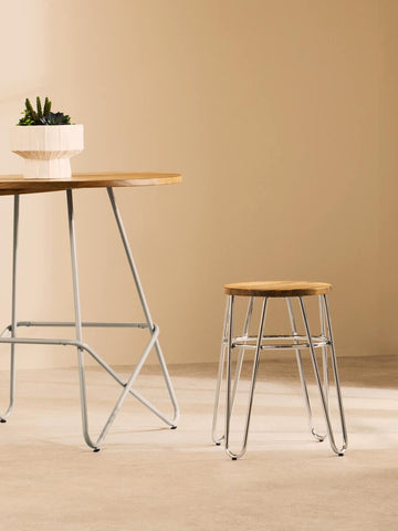 A minimalist stool featuring a round wooden top supported by sleek hairpin legs, positioned next to a matching table with a small potted plant on top, set against a soft, neutral background