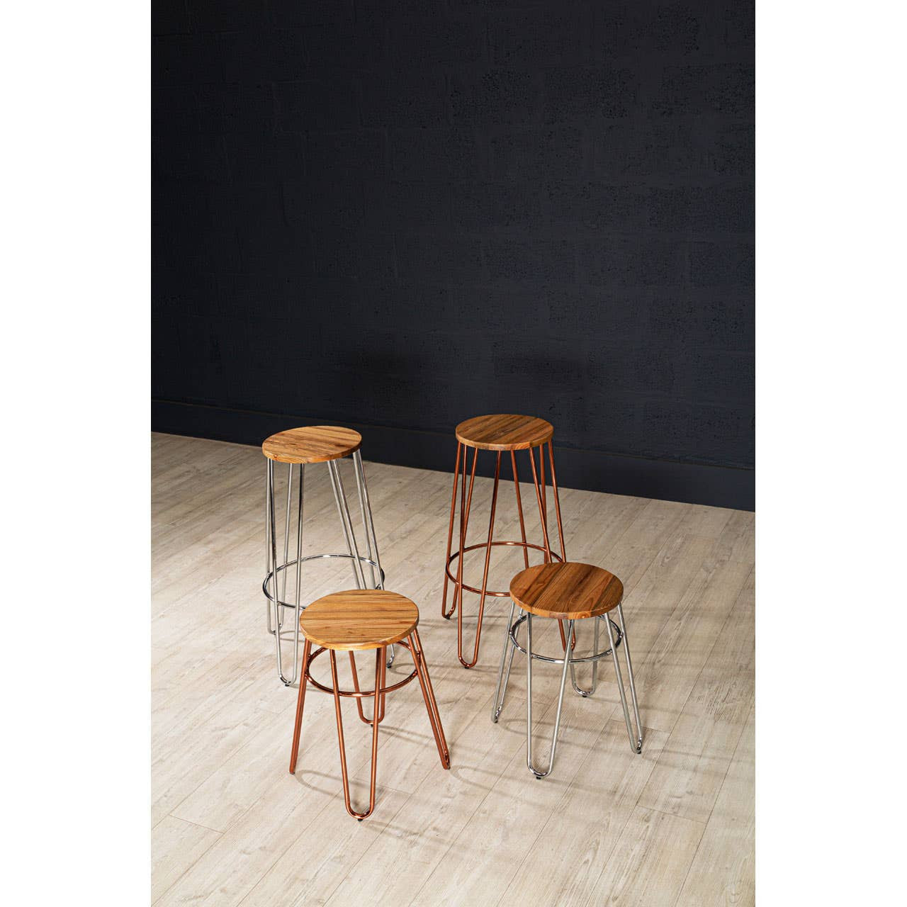 Four stools with wooden tops and hairpin metal legs are arranged on a light-colored floor against a dark wall. The stools vary in height, showcasing a modern design with a minimalist aesthetic