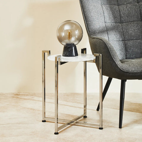 A white marble side table with a sleek chrome base, positioned next to a modern gray armchair. A decorative lamp sits on top of the table, casting a warm glow, with a light-colored wall and floor in the background