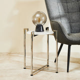 A white marble side table with a sleek chrome base, positioned next to a modern gray armchair. A decorative lamp sits on top of the table, casting a warm glow, with a light-colored wall and floor in the background