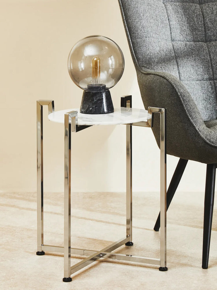 White marble side table with a sleek chrome base, featuring a minimalist design. A decorative globe lamp sits on top, and a modern chair is partially visible in the background. The floor is light-colored, enhancing the contemporary aesthetic of the scene