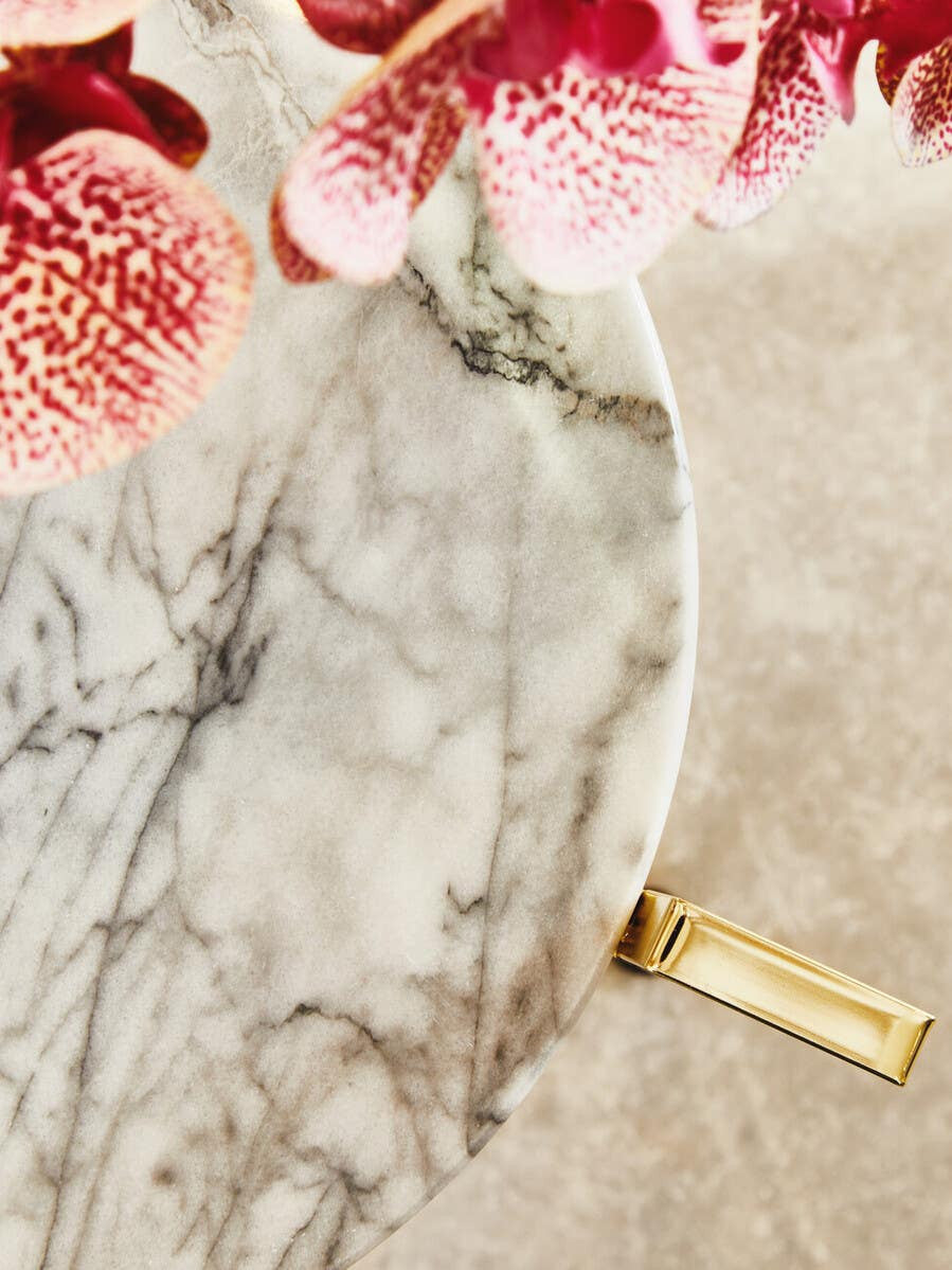 White marble side table featuring a smooth, rounded top with subtle gray veining, complemented by a warm gold base. The table is partially surrounded by pink flowers, adding a touch of color to the scene