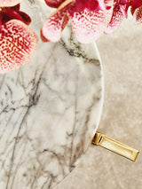 White marble side table featuring a smooth, rounded top with subtle gray veining, complemented by a warm gold base. The table is partially surrounded by pink flowers, adding a touch of color to the scene
