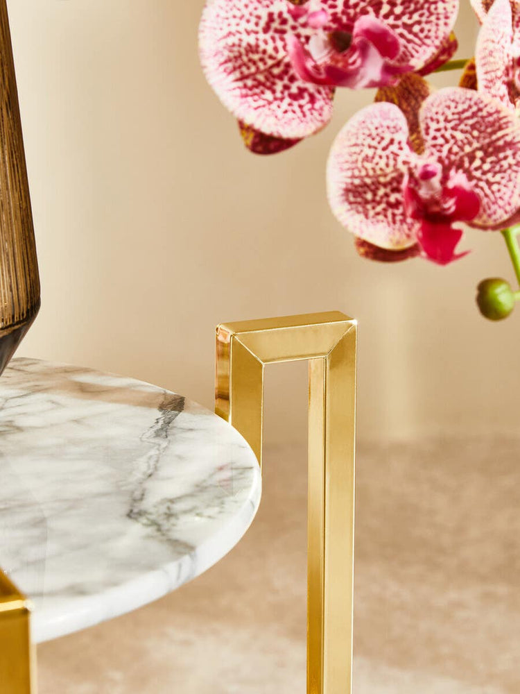 A white marble side table featuring a warm gold metal base, with a smooth circular top and sleek, modern legs. In the background, delicate pink orchids add a touch of color and elegance to the scene. The surface beneath the table is a light, neutral tone, enhancing the overall aesthetic
