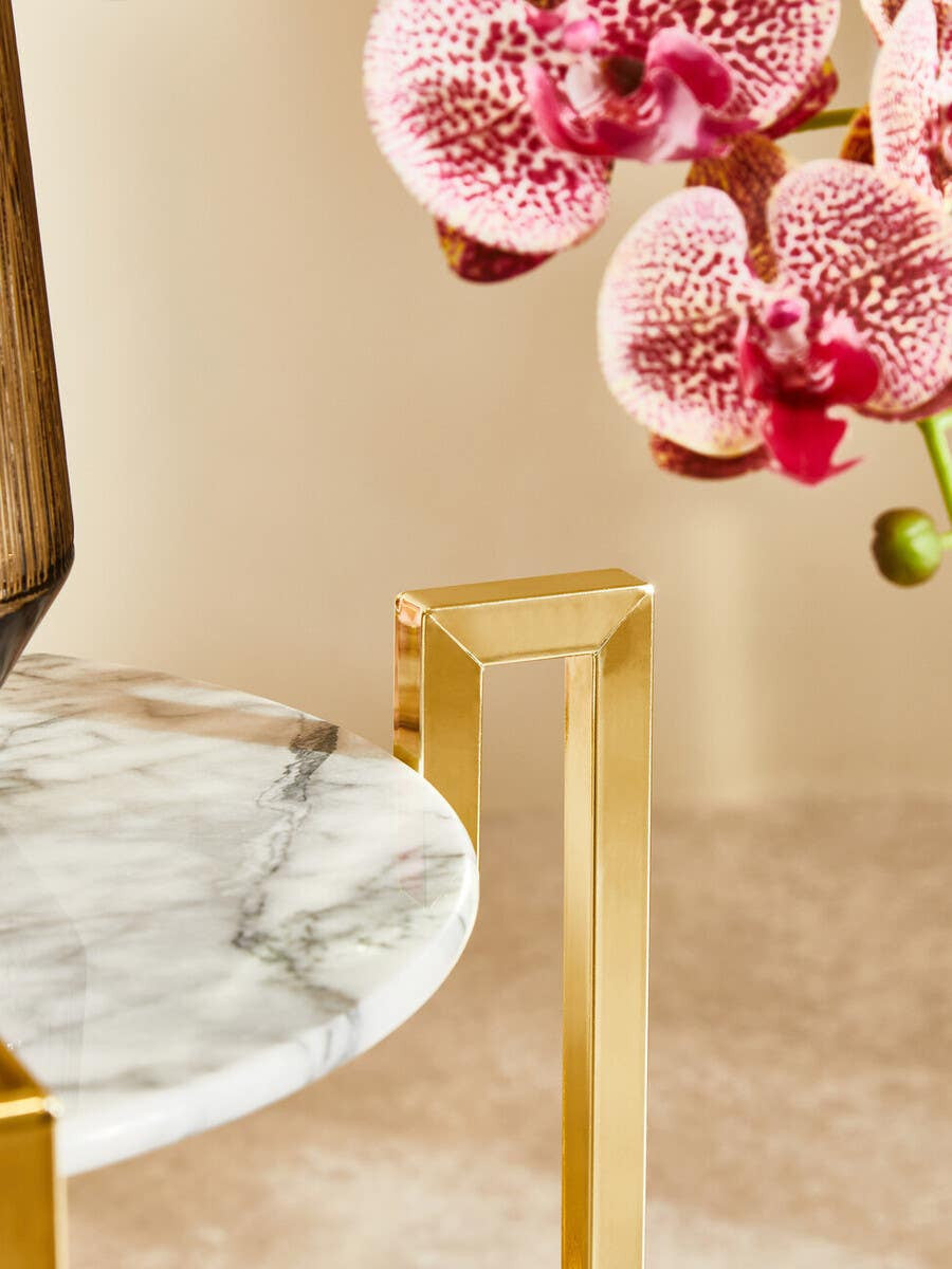 A white marble side table featuring a warm gold metal base, with a smooth circular top and sleek, modern legs. In the background, delicate pink orchids add a touch of color and elegance to the scene. The surface beneath the table is a light, neutral tone, enhancing the overall aesthetic