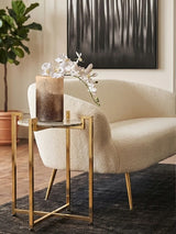 A white marble side table with a warm gold base is positioned next to a cream-colored curved armchair. The table holds a decorative glass vase filled with dried flowers, and a dark area rug is visible beneath the furniture. In the background, there is a large piece of abstract wall art and a potted plant