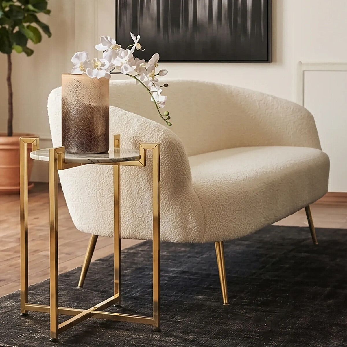 A white marble side table with a warm gold base, positioned next to a light-colored curved sofa. The table features a round top and slender legs, complementing the modern decor. A decorative vase with white flowers sits on the table, enhancing the elegant aesthetic of the room. The background includes a dark rug and a piece of artwork on the wall