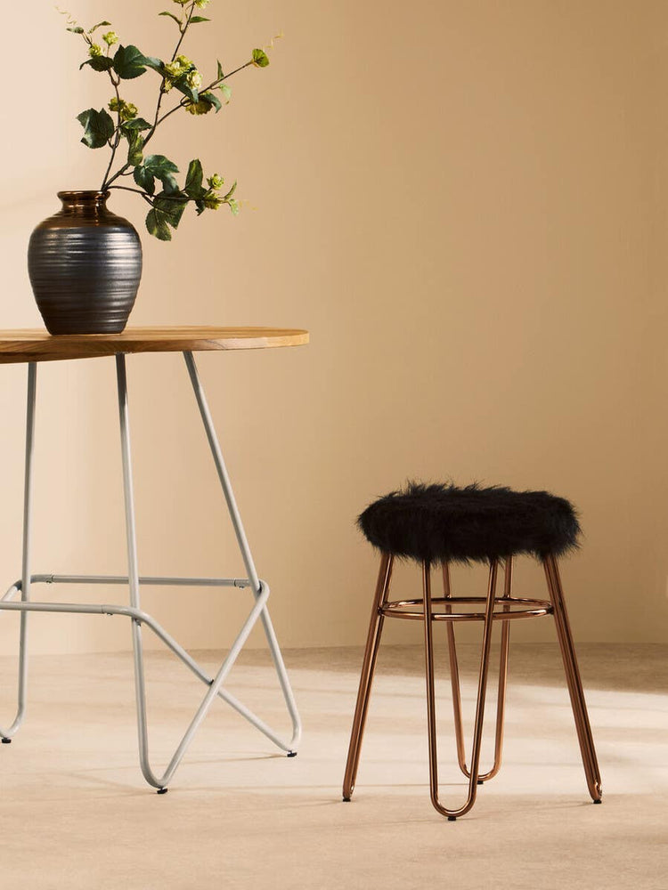 Faux fur stool with a rose gold finish, featuring a plush black seat and slender metal legs, positioned next to a wooden table with a vase of flowers against a neutral background