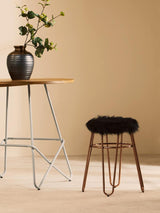 Faux fur stool with a rose gold finish, featuring a plush black seat and slender metal legs, positioned next to a wooden table with a vase of flowers against a neutral background
