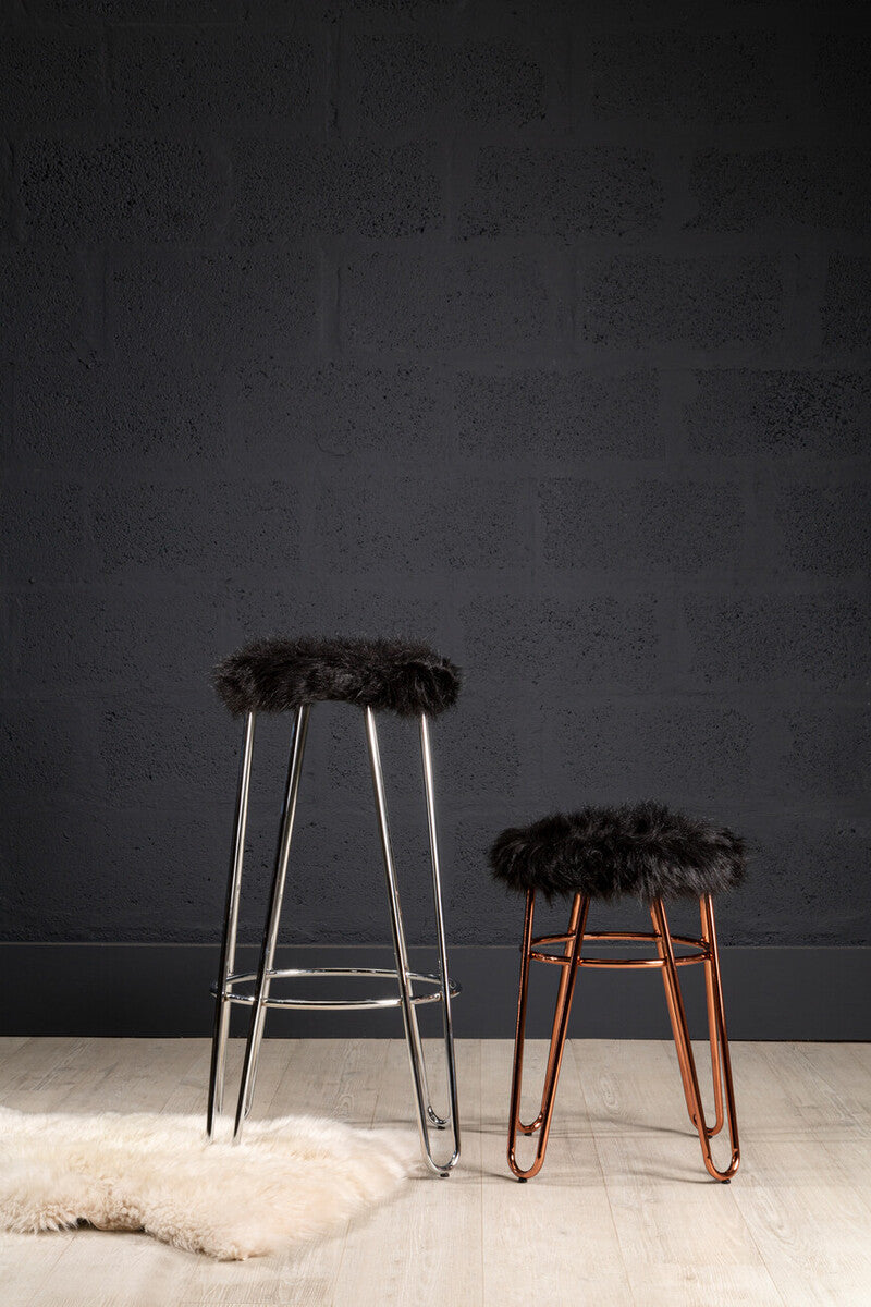Two stools are displayed against a dark wall. The taller stool features a faux fur seat with a rose gold finish on the legs, while the shorter stool has a similar design but with a darker wood finish. A soft, light-colored rug is positioned on the floor beneath them