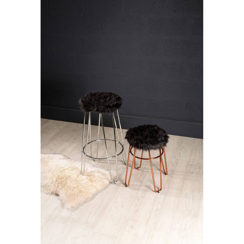 A faux fur stool with a rose gold finish base, featuring a plush black seat, positioned on a light wooden floor against a dark wall. Another similar stool is visible in the background