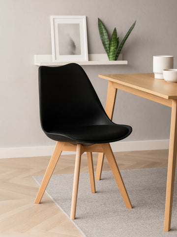 A sleek black chair with a curved seat and a soft cushion, supported by light beech wood legs, positioned next to a wooden table in a modern interior setting. A small potted plant and a decorative frame are visible on a shelf in the background, with a neutral-colored rug beneath the chair