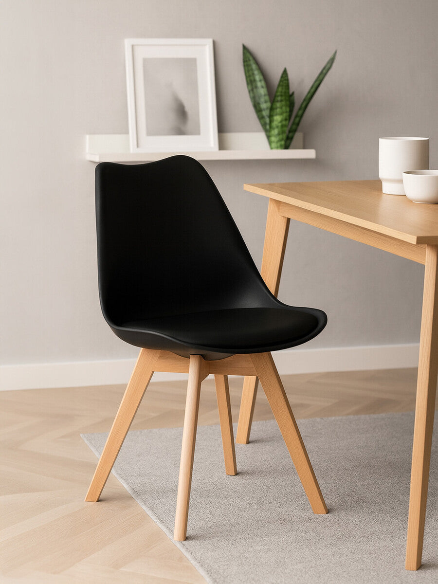 A sleek black chair with a curved seat and a soft cushion, supported by light beech wood legs, positioned next to a wooden table in a modern interior setting. A small potted plant and a decorative frame are visible on a shelf in the background, with a neutral-colored rug beneath the chair