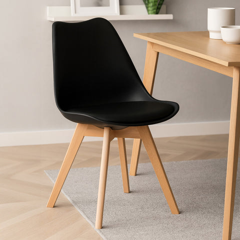 A sleek black chair with a curved backrest and a cushioned seat, supported by light beech wood legs. The chair is positioned next to a wooden table on a light-colored rug, with a neutral wall and a window in the background